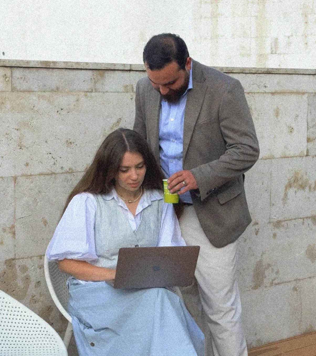 A woman sits on a chair holding a laptop, looking up at a man standing next to her who is holding a cup.