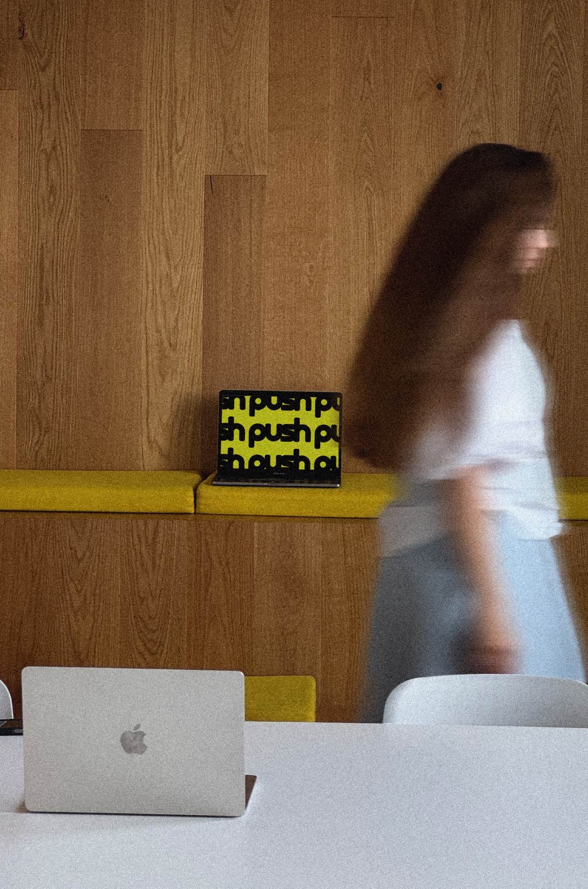 Blurred person walking past a wooden wall with a yellow bench. A laptop with a yellow "push" pattern is on the bench, and a closed MacBook is on a white table in the foreground.