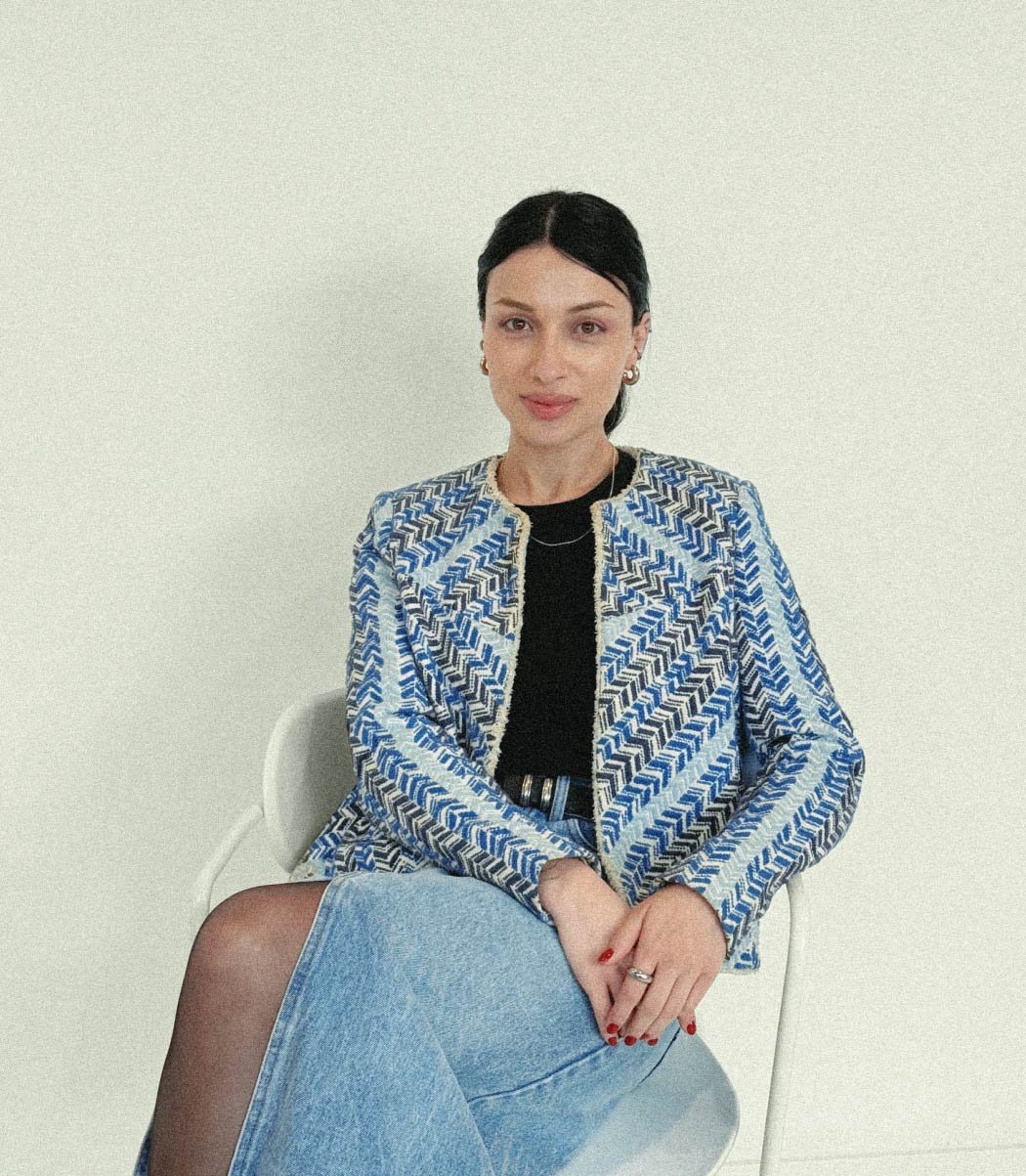 A woman with dark hair sits on a white chair, wearing a blue patterned jacket, black top, and denim skirt, with her hands resting on her lap - portrait of Marina.