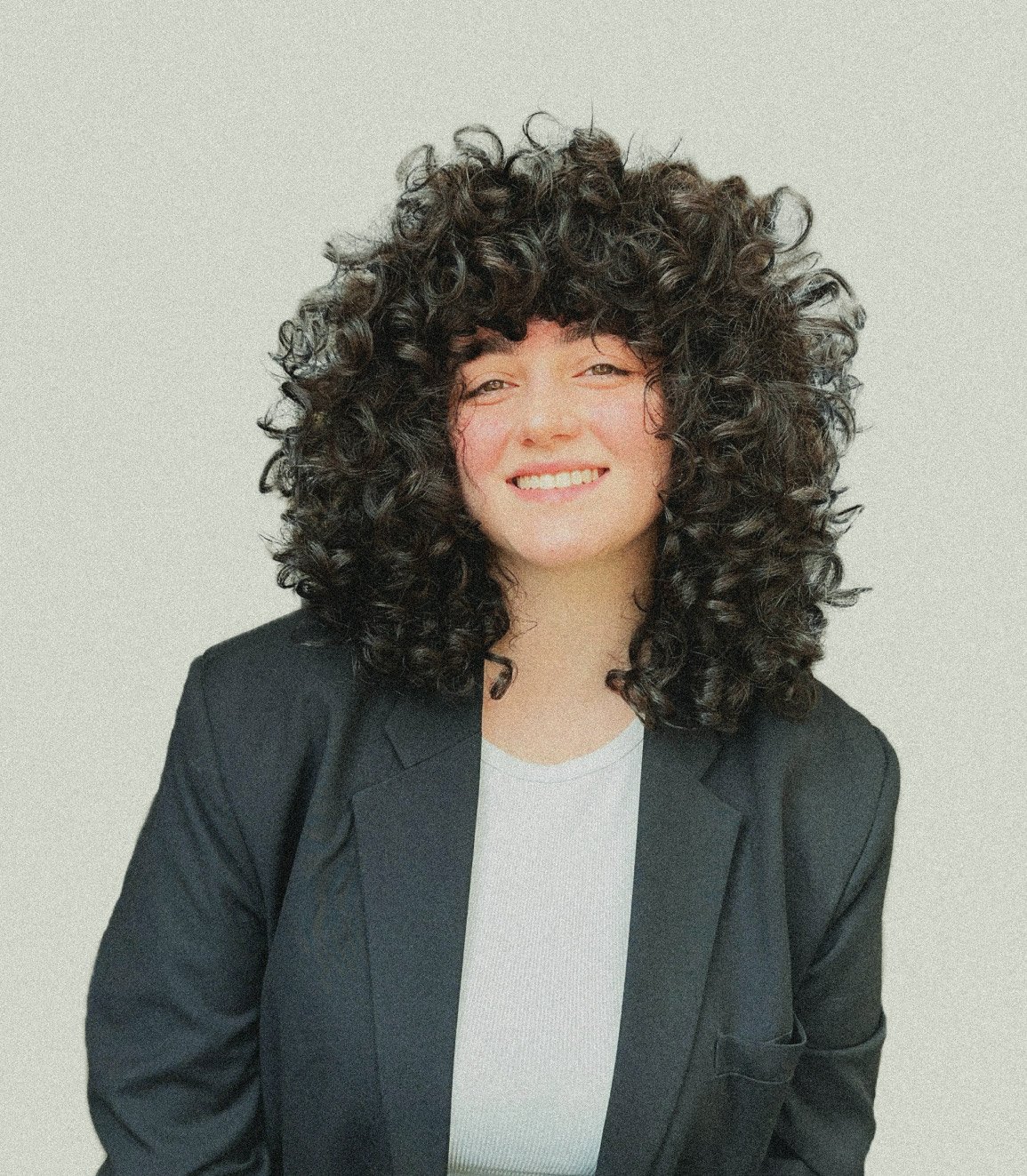 A person with curly hair and black blazer over a white shirt smiles at the camera.