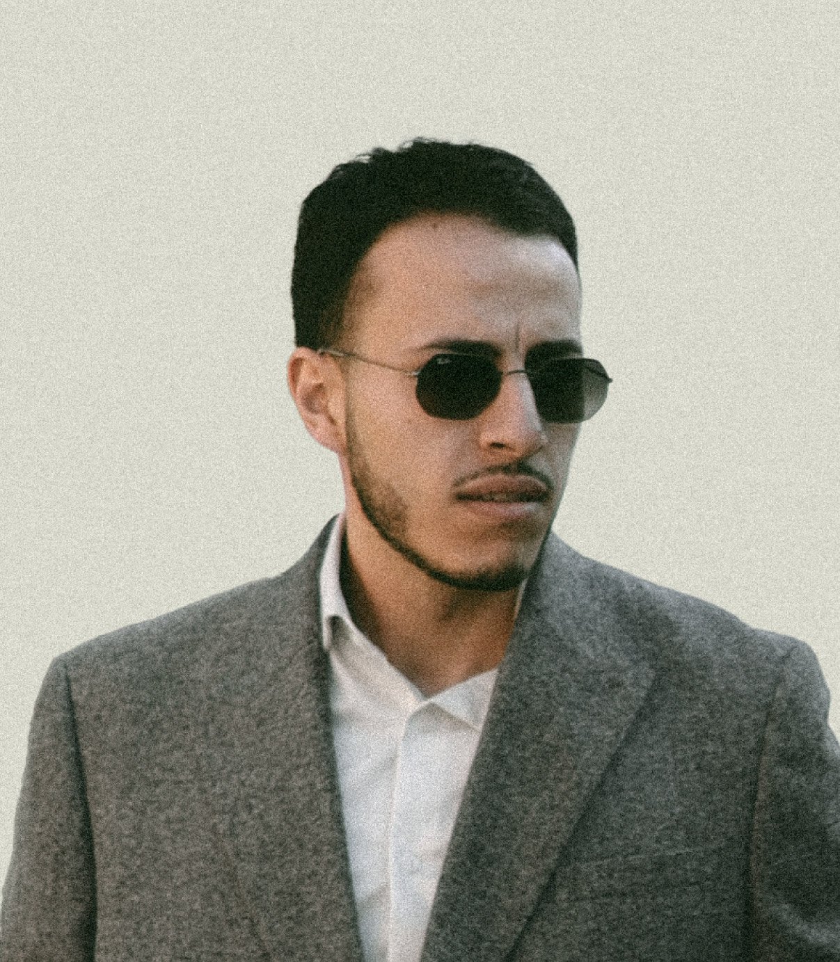 A man wearing dark sunglasses and a gray suit jacket over a white shirt looks to the side
